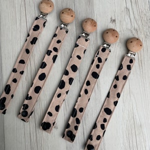 May include: Five pacifier clips. The tan fabric has black polka dots. Each clip has a wooden bead, a metal clip, and a black snap closure. The clips are arranged diagonally on a light wood-grain surface.
