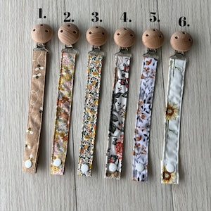 May include: Six pacifier clips with different fabric patterns. The clips are made of wood and have a metal clasp. The fabric patterns include bees, floral, and sunflowers.