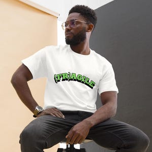 May include: A white t-shirt with the word "FRAGILE" in green and black lettering. The t-shirt is being worn by a person sitting on a black stool. The person is wearing black jeans and white trainers.