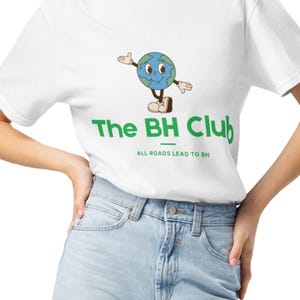 Può includere: T-shirt bianco con grafica verde e blu di un globo terrestre con braccia e gambe. Il testo "The BH Club" è in verde, con "ALL ROADS LEAD TO BH" sotto. La maglietta è abbinata a jeans azzurri.
