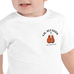 May include: White toddler t-shirt featuring the text "LIL SLUGGER" above a pair of orange boxing gloves. The shirt also has the text "000 STUDIO GOODS" below the gloves.