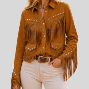 Puede incluir: Camisa de ante marrón con flecos en el pecho, las mangas y los bolsillos. La camisa presenta detalles de tachuelas plateadas y una botonadura frontal. Se ve un cinturón de cuero marrón con hebilla plateada.