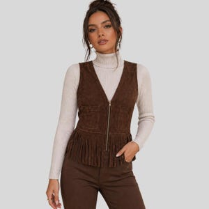 Puede incluir: Un chaleco de ante marrón con cuello en V, cierre de cremallera y flecos en el dobladillo. El chaleco tiene dos pequeños bolsillos y se lleva sobre un cuello alto color crema. La modelo también lleva pantalones marrones.