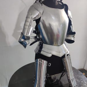 May include: A polished steel suit of armor featuring a breastplate, articulated shoulder pauldrons, upper arm defenses, and lower leg greaves. The metallic components are secured with black leather straps and rivets, displayed on a black mannequin torso.