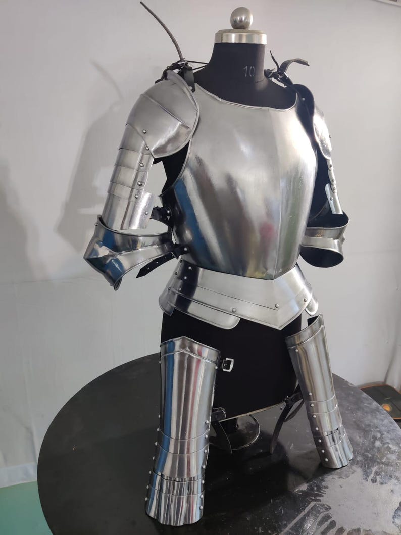 May include: A polished steel suit of armor featuring a breastplate, articulated shoulder pauldrons, upper arm defenses, and lower leg greaves. The metallic components are secured with black leather straps and rivets, displayed on a black mannequin torso.