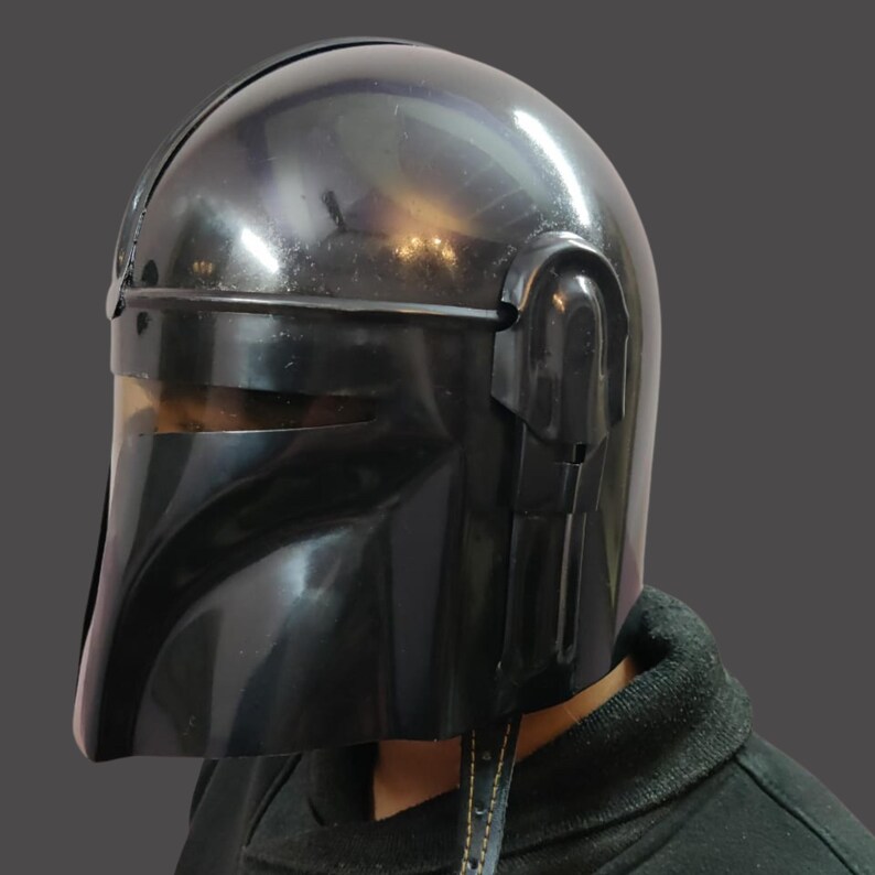 Puede incluir: Un casco negro brillante con visera y orejeras. El casco tiene una superficie lisa y reflectante y un dise&ntilde;o angular distintivo. El casco es un art&iacute;culo de cosplay.