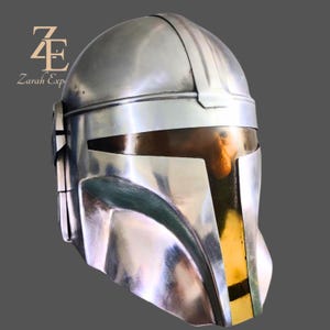 Puede incluir: Un casco mandaloriano plateado con una superficie reflectante. El casco presenta una visera con una secci&oacute;n dorada y una abertura rectangular oscura. El casco es una r&eacute;plica del que usa el personaje de la serie Star Wars.