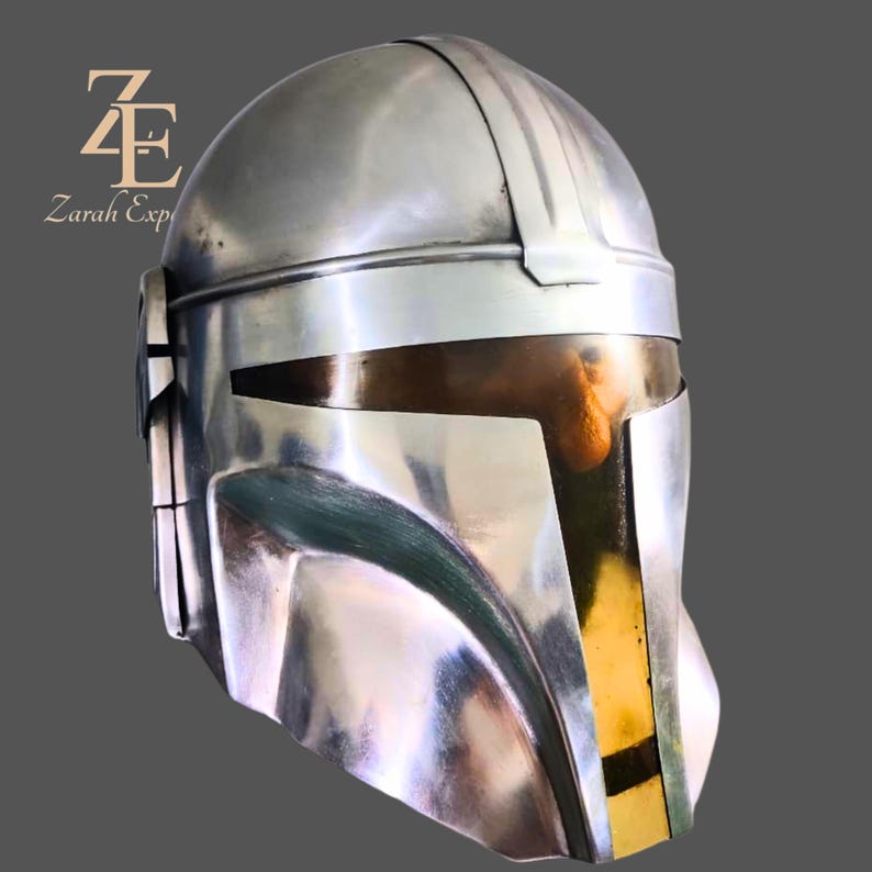 Puede incluir: Un casco mandaloriano plateado con una superficie reflectante. El casco presenta una visera con una secci&oacute;n dorada y una abertura rectangular oscura. El casco es una r&eacute;plica del que usa el personaje de la serie Star Wars.