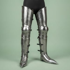 May include: A pair of polished steel leg armor pieces featuring articulated knee caps, thigh guards, and pointed sabatons. The armor is secured with black leather straps and buckles along the outer sides, covering the legs from thigh to foot.