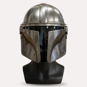 Puede incluir: Un casco plateado de Mandaloriano con una visera oscura, exhibido en una cabeza de maniqu&iacute; negra. El casco tiene una apariencia desgastada con detalles visibles y una cresta vertical central.