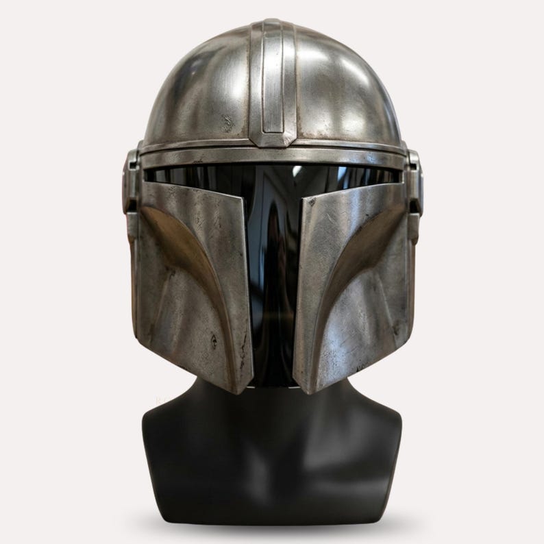 Puede incluir: Un casco plateado de Mandaloriano con una visera oscura, exhibido en una cabeza de maniqu&iacute; negra. El casco tiene una apariencia desgastada con detalles visibles y una cresta vertical central.