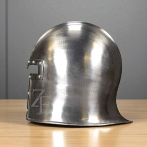 K&ouml;nnte beinhalten: Ein polierter, silberfarbener Metallhelm mit abgerundeter Oberseite und einem ausgestellten Nackenschutz. Der Helm hat einen rechteckigen Ausschnitt an der Seite und ein stilisiertes Logo. Die Oberfl&auml;che reflektiert das Licht und hebt seine glatte Textur hervor. Der Helm ist wahrscheinlich f&uuml;r historische Nachstellungen oder Ausstellungen.