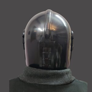 Puede incluir: Un casco negro con un acabado brillante, con una cresta central y detalles rectangulares en la parte trasera. El casco se lleva sobre un cuello alto negro.
