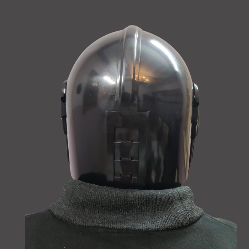 Puede incluir: Un casco negro con un acabado brillante, con una cresta central y detalles rectangulares en la parte trasera. El casco se lleva sobre un cuello alto negro.