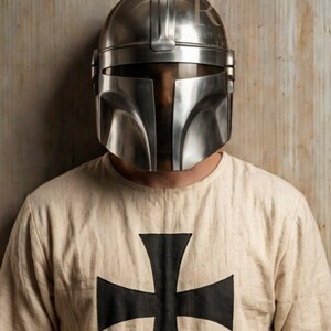 Puede incluir: Un casco plateado de estilo mandaloriano con una visera en forma de T. El casco se lleva sobre una camisa color crema con una gran cruz negra en la parte delantera. La camisa tiene cuello redondo.