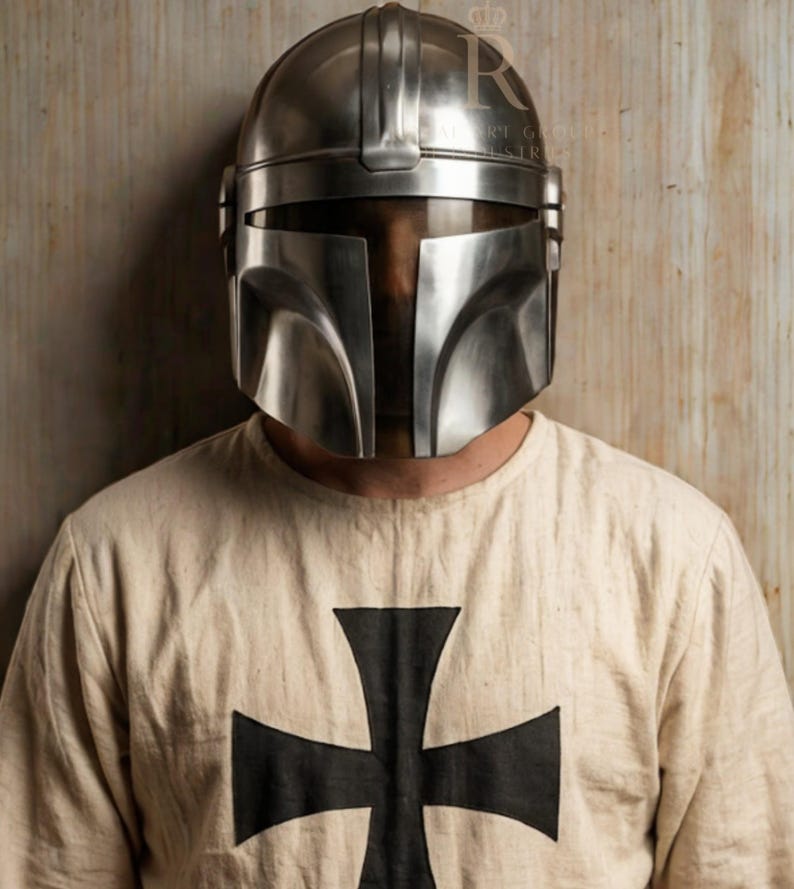 Puede incluir: Un casco plateado de estilo mandaloriano con una visera en forma de T. El casco se lleva sobre una camisa color crema con una gran cruz negra en la parte delantera. La camisa tiene cuello redondo.