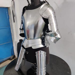May include: A polished steel suit of armor featuring a breastplate, pauldrons, and articulated arm and leg plating. The metallic components are secured with rivets and leather straps, displayed on a dark mannequin torso to show the structural design.