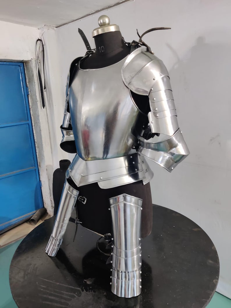 May include: A polished steel suit of armor featuring a breastplate, pauldrons, and articulated arm and leg plating. The metallic components are secured with rivets and leather straps, displayed on a dark mannequin torso to show the structural design.