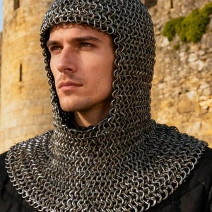 May include: A close-up of a chainmail coif, a type of medieval head covering, worn over a black quilted garment. The chainmail is made of small, interlocking metal rings. The background features a stone castle.