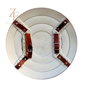 May include: A silver-colored, round shield with a series of concentric circles. The shield has brown leather straps and buckles on the back for securing it to the arm. The shield is a prop or decorative item.
