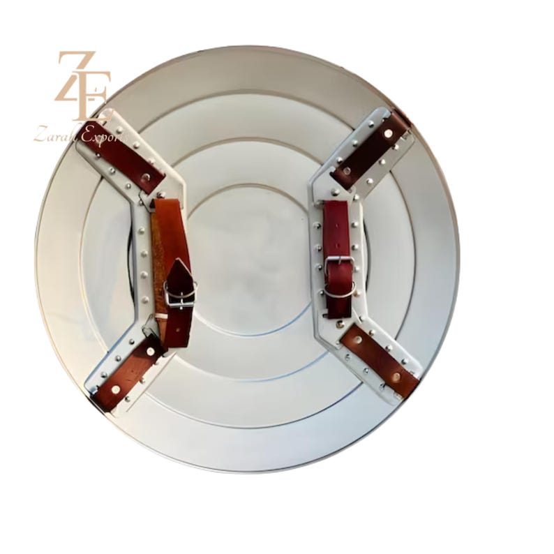 May include: A silver-colored, round shield with a series of concentric circles. The shield has brown leather straps and buckles on the back for securing it to the arm. The shield is a prop or decorative item.
