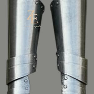 May include: A pair of polished steel greaves featuring a multi-plate construction with visible rivets along the edges. The metal armor pieces are curved to fit the lower leg and include the logo ZE Z Exports printed in gold on the left piece.