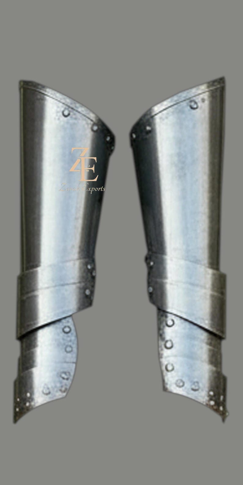 May include: A pair of polished steel greaves featuring a multi-plate construction with visible rivets along the edges. The metal armor pieces are curved to fit the lower leg and include the logo ZE Z Exports printed in gold on the left piece.