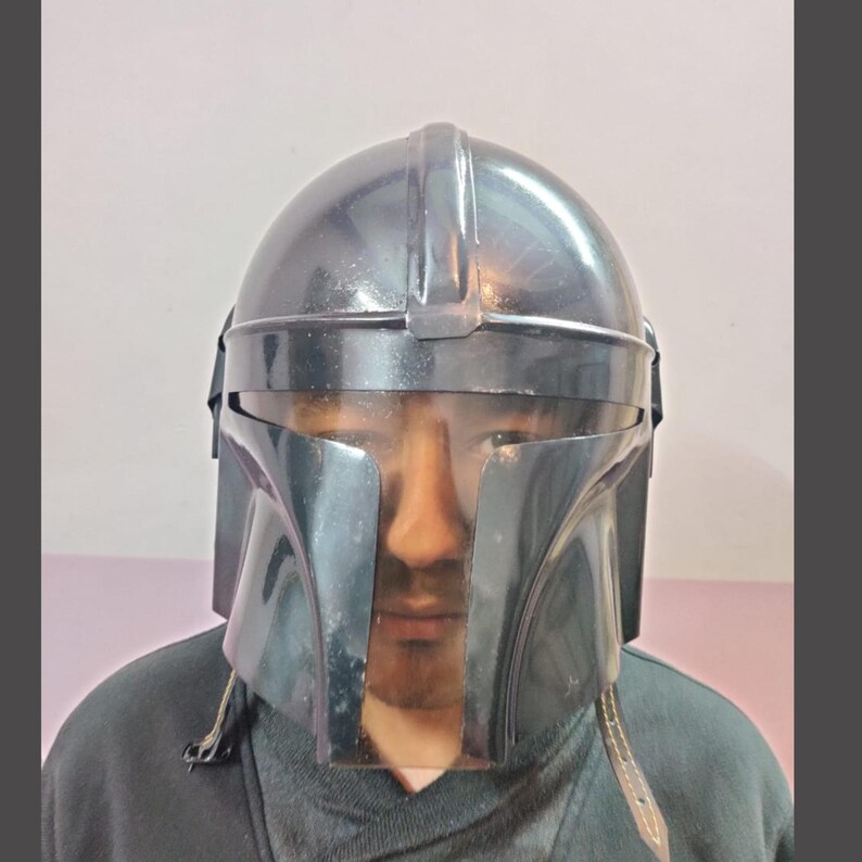 Puede incluir: Un casco mandaloriano gris oscuro con una visera transparente. El casco tiene una forma distintiva con una parte superior redondeada y rasgos angulares. La visera es transparente y el casco parece estar hecho de un material duradero.
