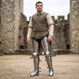 May include: A quilted olive green vest worn over a beige long-sleeved shirt, paired with polished steel leg armor. The metal greaves and poleyns feature articulated plates and black leather straps, covering the legs from the knee to the foot.