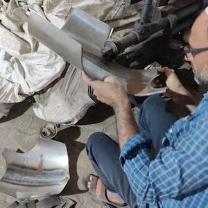 May include: A person uses a metalworking machine to shape a curved piece of silver-colored sheet metal. The metal features visible weld marks and a smooth, reflective surface. Additional pre-cut metal components are scattered nearby on the floor.