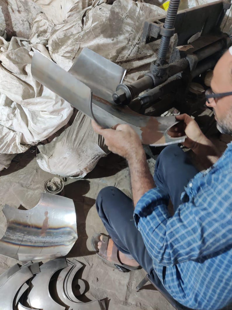 May include: A person uses a metalworking machine to shape a curved piece of silver-colored sheet metal. The metal features visible weld marks and a smooth, reflective surface. Additional pre-cut metal components are scattered nearby on the floor.