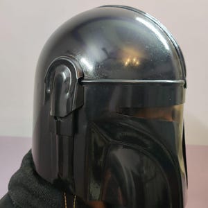 Puede incluir: Un casco negro brillante con un dise&ntilde;o distintivo, con una parte superior redondeada y una visera angular. El casco tiene una superficie lisa y reflectante y probablemente est&aacute; hecho de un material duradero. El dise&ntilde;o recuerda a un personaje tem&aacute;tico espacial.
