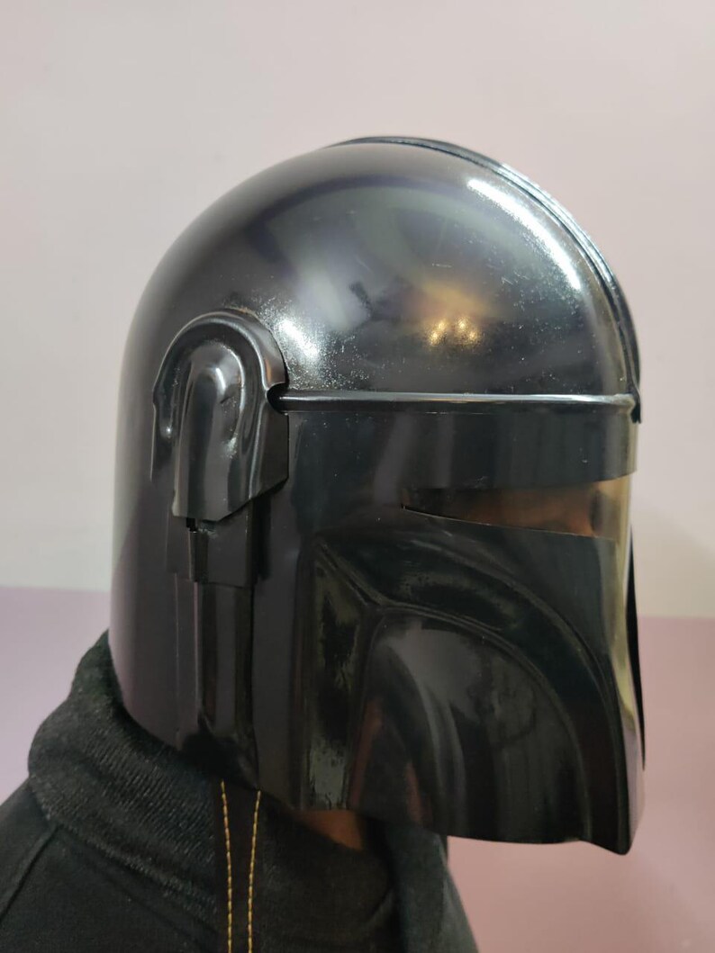 Puede incluir: Un casco negro brillante con un dise&ntilde;o distintivo, con una parte superior redondeada y una visera angular. El casco tiene una superficie lisa y reflectante y probablemente est&aacute; hecho de un material duradero. El dise&ntilde;o recuerda a un personaje tem&aacute;tico espacial.