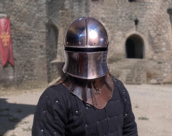 May include: A polished steel helmet with a rounded dome and a horizontal visor slit, featuring small gold-toned rivets along the brow. The helmet is attached to a brown leather gorget with a metal buckle, worn over a black quilted long-sleeve tunic.