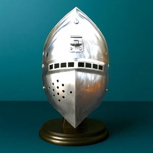 Puede incluir: Un yelmo de caballero plateado pulido con una parte superior puntiaguda y una visera con orificios de ventilación rectangulares y circulares. El yelmo está montado sobre una base de madera oscura, sobre un fondo verde azulado.