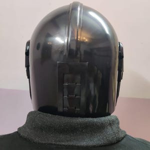 Puede incluir: Un casco negro con la parte superior redondeada y paneles laterales. El casco tiene un acabado brillante y un panel rectangular en la parte trasera con una serie de crestas horizontales. La persona lleva un cuello alto negro.