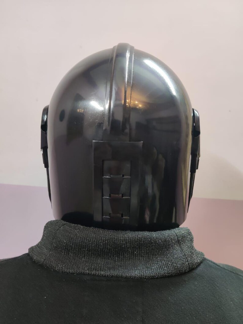 Puede incluir: Un casco negro con la parte superior redondeada y paneles laterales. El casco tiene un acabado brillante y un panel rectangular en la parte trasera con una serie de crestas horizontales. La persona lleva un cuello alto negro.