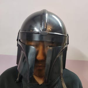 Puede incluir: Un casco negro mandaloriano con una visera transparente. El casco tiene una parte superior redondeada y rasgos angulares, con una superficie oscura y reflectante. La visera es transparente y el casco parece estar hecho de un material duradero.