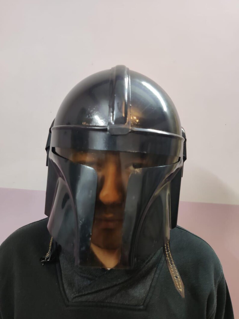 Puede incluir: Un casco negro mandaloriano con una visera transparente. El casco tiene una parte superior redondeada y rasgos angulares, con una superficie oscura y reflectante. La visera es transparente y el casco parece estar hecho de un material duradero.