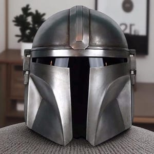 Puede incluir: Un casco mandaloriano plateado y verde oscuro. El casco tiene una visera en forma de T y una parte superior redondeada con una cresta central. El dise&ntilde;o presenta placas angulares en las mejillas y un acabado desgastado, lo que sugiere una est&eacute;tica desgastada por la batalla.