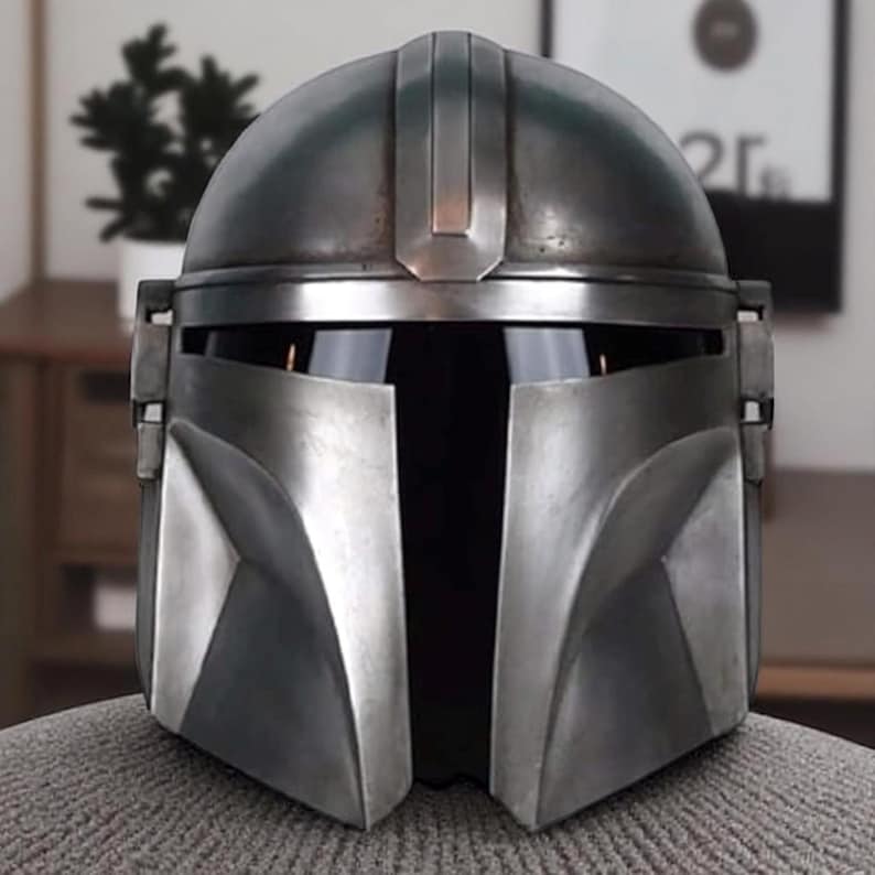 Puede incluir: Un casco mandaloriano plateado y verde oscuro. El casco tiene una visera en forma de T y una parte superior redondeada con una cresta central. El dise&ntilde;o presenta placas angulares en las mejillas y un acabado desgastado, lo que sugiere una est&eacute;tica desgastada por la batalla.