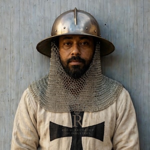 May include: A metallic kettle hat helmet with a central ridge and rivets, paired with a silver chainmail aventail. The garment is a cream-colored long-sleeve tunic featuring a large black cross and the text Royal Art Group of Industries.