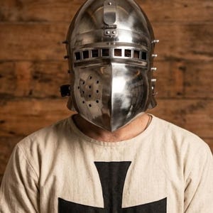 May include: A shiny, silver knight's helmet with a visor and rivet details. The person is wearing a cream-colored shirt with a large black cross on the front. The background is a wooden wall.