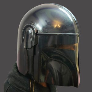 Puede incluir: Un casco negro mandaloriano con un acabado brillante. El casco presenta una parte superior redondeada, una visera y detalles laterales distintivos. El dise&ntilde;o es reminiscente de un personaje de tem&aacute;tica espacial, adecuado para cosplay o exhibici&oacute;n.