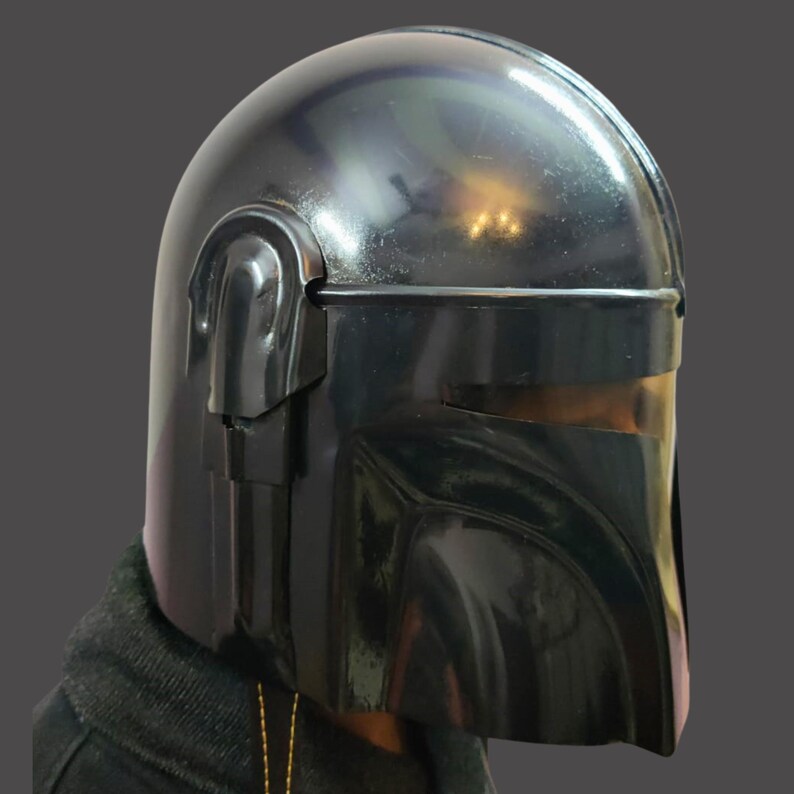 Puede incluir: Un casco negro mandaloriano con un acabado brillante. El casco presenta una parte superior redondeada, una visera y detalles laterales distintivos. El dise&ntilde;o es reminiscente de un personaje de tem&aacute;tica espacial, adecuado para cosplay o exhibici&oacute;n.