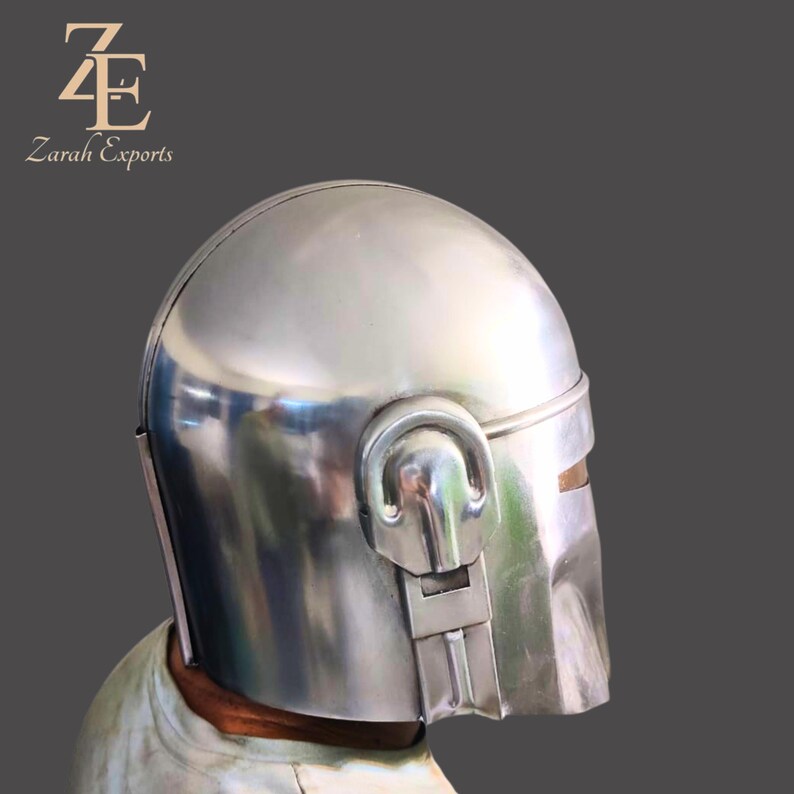 Puede incluir: Un casco de metal plateado pulido con una parte superior redondeada y una visera. El casco tiene una secci&oacute;n con bisagras en el lateral y una abertura rectangular para la visera. El logotipo "Zarah Exports" es visible en la esquina superior izquierda.