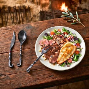 May include: A set of hand-forged black metal cutlery including a knife, a spoon, and a fork. The fork is placed on a white ceramic plate containing a meal of sliced grilled meat, a fish fillet, and a fresh green salad with cherry tomatoes.