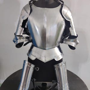 May include: A polished steel suit of armor displayed on a mannequin. The set includes a breastplate, shoulder pauldrons, arm guards, and leg greaves, all featuring a reflective metallic finish and secured with black leather straps and metal buckles.