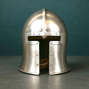 K&ouml;nnte beinhalten: Ein polierter silberner mittelalterlicher Helm mit spitzem Oberteil und rechteckigen Augen&ouml;ffnungen. Nieten sichern die Frontplatte und die Seitenteile. Ein brauner Lederriemen ist im Helm sichtbar. Der Helm wird vor einem dunkelblauen Hintergrund gezeigt.