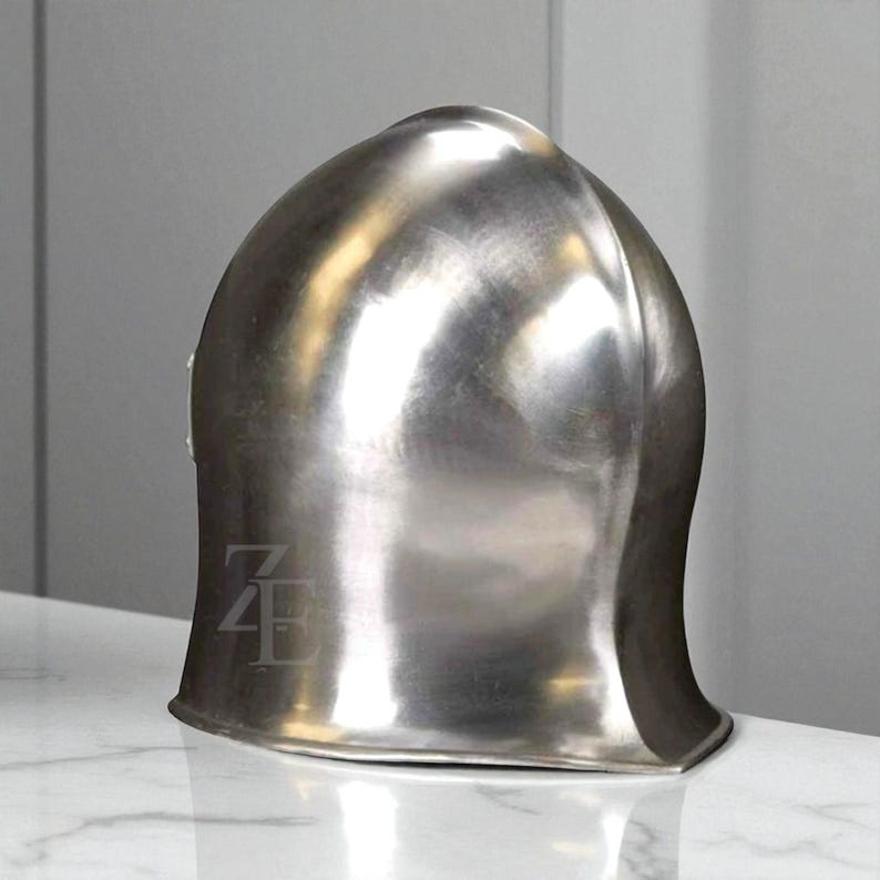 K&ouml;nnte beinhalten: Ein polierter silberner Helm mit abgerundeter Oberseite und ausgestelltem Boden. Der Helm hat eine glatte, reflektierende Oberfl&auml;che, wahrscheinlich aus Metall. Das Design deutet auf ein historisches oder mittelalterliches Thema hin.